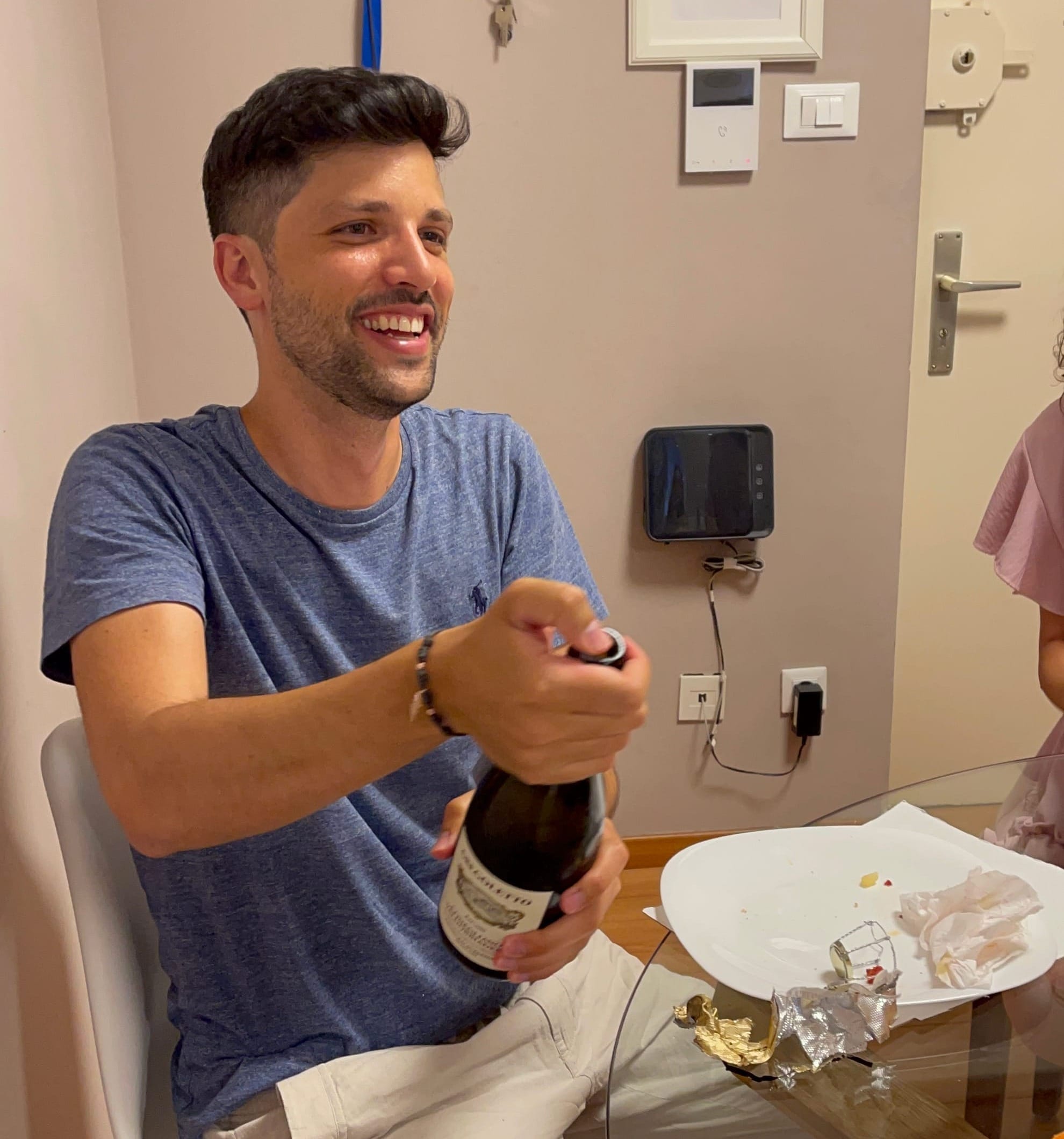Photo of Marco sitting in a kitchen, wearing a blue t-shirt and smiling while opening a bottle of champagne, capturing a moment of celebration.